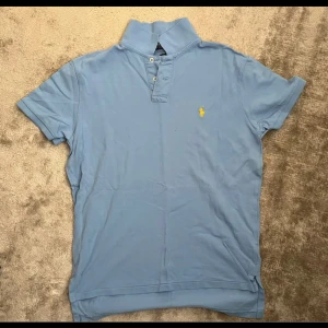Ljusblå pikétröja från Polo Ralph Lauren - Snygg ljusblå pikétröja från Polo Ralph Lauren med klassisk krage, korta ärmar och gul broderad logga på bröstet. Tröjan är i bomull och har knäppning med två knappar framtill. Perfekt för en clean och stilren look.