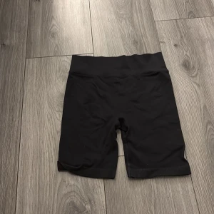 Svarta ribbade cykelshorts New Apparel - Svarta cykelshorts från New Apparel i ribbat material. De är högmidjade och har en tight passform som sitter skönt mot kroppen. Tillverkade i stretchig polyester och spandex, perfekta för träning eller chill. Snygg minimalistisk look utan synliga detaljer.