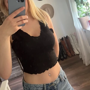 Basic Svart ribbad croppad linnetopp - Säljer en svart croppad linnetopp med ribbad struktur och vågiga kanter vid halsringning och nederkant. Toppen är ärmlös och har en snygg v-ringning. Perfekt att matcha med jeans eller kjol för en trendig look.