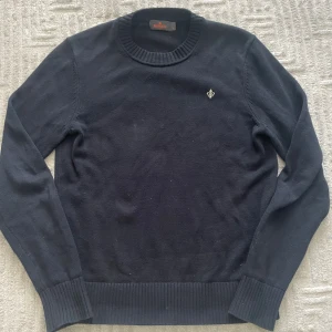 Mörkblå stickad tröja från Morris - Klassisk mörkblå stickad tröja från Morris med rund halsringning och ribbade muddar. Tröjan har en liten broderad logga på bröstet och är perfekt för dig som gillar stilrena och tidlösa plagg. Priset är inte hugget i sten! Vid fler frågor eller funderingar tveka inte på att skriva!