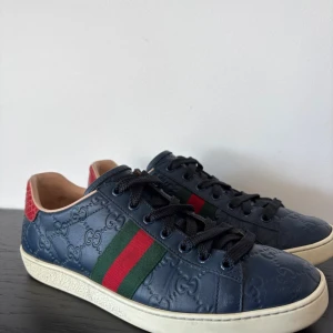 Gucci sneakers i blått skinn - (INGA BYTEN) Säljer ett par exklusiva Gucci Ace sneakers i marinblå läder med präglad branding. Skorna har den klassiska gröna och röda sidan, svarta skosnören, vit sula och en röd-grön texturerad hälpatch. De är i mycket bra skick och ger en stilren och lyxig look som passar både till vardags och fest. Storlek [ange storlek]. Perfekta för dig som vill ha en tidlös design med hög kvalitet! 