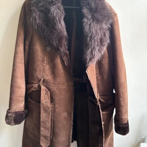Brun pälsfodrad mockakappa - Säljer en brun lång kappa i mocka med bred pälskrage och pälsdetaljer vid ärmslut. Kappan har stora fickor, bälte i midjan och knäppning framtill. Perfekt för dig som vill ha en retro och riktigt varm look.