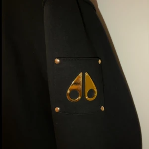 Svart hoodie Moose Knuckles XL - Svart hoodie från Moose Knuckles i storlek XL med guldfärgade detaljer och dragkedja. Tröjan har huva, fickor framtill och ett snyggt guldemblem på ärmen. Insidan är fodrad med mjukt material för extra komfort och värme.