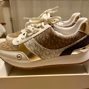 *NY*Michael Kors sneakers Andi stl 6,5 (stl 36/36,5) Presenttips  - Nya oanvända i kartong med prislapp. Ord pris: 2800 sek Outlet: 1860 sek  Pris nu: 550 sek.        Innermått: 22,5 cm • Trainer • Logo-print canvas • Lining: polyester • Sole: rubber • Round toe. • Lace-up fastening. • Imported. Snygga sneakers från Michael Kors med MK-logga i monogrammönster. Skorna har detaljer i guld, beige och vitt, samt vita snören och guldmetallic på häl och sidor. Ovandelen är i textil och syntet med skinnliknande partier. Perfekta för dig som gillar lyxig streetstyle. 