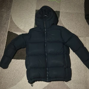 Mörk blå pufferjacka från Peak Performance - Säljer en svart pufferjacka från Peak Performance med huva och dragkedja. Jackan är tjockt vadderad och har en clean look med diskret logga på bröstet. Perfekt för kalla dagar och riktigt snygg till streetwear-stilen.