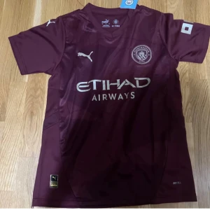 Vinröd Manchester City fotbollströja Puma - Snygg vinröd Manchester City fotbollströja från Puma med Etihad Airways-tryck på bröstet. Klassisk passform med korta ärmar och rund hals. Tillverkad i lätt och ventilerande material, perfekt för match eller träning.
