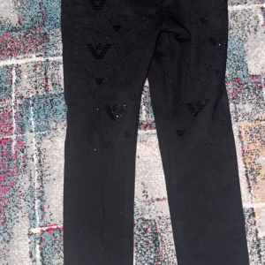 Svarta jeansbyxor från Armani - Svarta jeansbyxor från Armani med raka ben och diskreta svarta loggor framtill. Byxorna har klassisk femficksdesign och är tillverkade i ett stretchigt bomullsmaterial. Snygga detaljer med små svarta nitar och metallmärke bak i midjan.