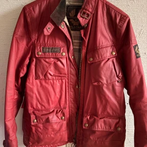 Röd Belstaff jacka med rutigt foder - Snygg röd jacka från Belstaff med flera fickor, dragkedja och tryckknappar. Jackan har ett rutigt, quiltat innerfoder i blått, grått och beige. Klassisk krage med spänne och detaljerade logopatchar. Perfekt för dig som gillar street och utility vibes.