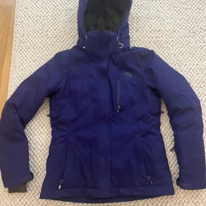 Blå skidjacka från The North Face - Snygg mörkblå/lila vindjacka från The North Face med huva och dragkedja framtill. Jackan har flera fickor med dragkedja och justerbara ärmslut. 