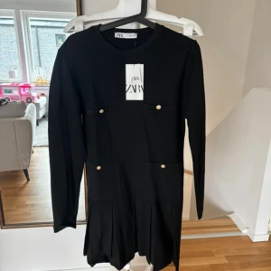 Svart kort klänning från Zara - Stilren svart klänning från Zara med lång ärm, rund halsringning och dekorativa guldknappar på fickor framtill. Kjolen har snygga veck för extra volym. Perfekt för dig som gillar klassisk men trendig look. Nypris 500kr