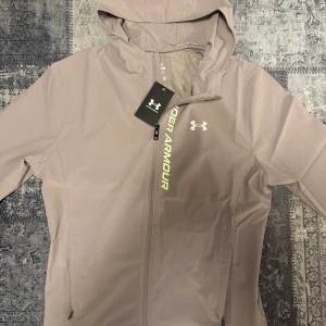 Grå vindjacka från Under Armour - Säljer en grå vindjacka från Under Armour med huva och dragkedja framtill. Jackan har logga på bröstet och är tillverkad i ett lätt syntetmaterial. Perfekt för blåsiga dagar och har snyggt tryck längs dragkedjan. Modellen är fitted och har meshfoder på insidan.