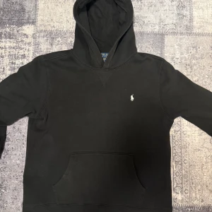 Svart hoodie från Polo Ralph Lauren - Svart hoodie från Polo Ralph Lauren med klassisk huva och känguruficka. Tröjan har en broderad vit logga på bröstet och är tillverkad i mjuk bomull. Perfekt för chill dagar och enkel att matcha med jeans eller joggers.