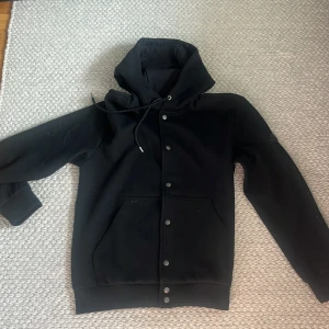 Svart hoodie från Moncler med knappar - Svart hoodie från Moncler med huva och snörning. Tröjan har tryckknappar framtill och stora fickor på magen. Materialet känns mjukt och lite tjockare, perfekt för kyligare dagar. Klassisk och stilren design som funkar till det mesta.