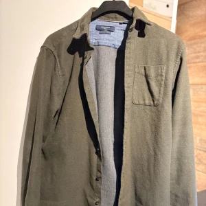 Olivgrön skjorta från Jack & Jones - Olivgrön skjorta från Jack & Jones med klassisk krage, bröstficka och knäppning framtill. Skjortan är långärmad och har en avslappnad passform, perfekt att bära öppen över en t-shirt. Tillverkad i mjuk bomull för en skön känsla.