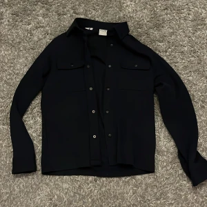 Svart overshirt från Selected Homme - Stilren svart overshirt från Selected Homme med klassisk krage, två bröstfickor med lock och knappar framtill. Perfekt att slänga över en t-shirt för en clean look. Långärmad och i ett mjukt, lite tjockare material. Nypris 600