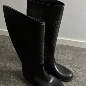 Svarta quiltade stövlar - Säljer ett par svarta stövlar med quiltad detalj på skaftet. De har rund tå och är tillverkade i syntetmaterial med en glansig finish nedtill och matt quiltad ovandel. Perfekta för regniga dagar och ger en stilren look.