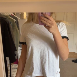 Vit t-shirt med spets från Zara - Vit t-shirt från Zara i bomull med korta ärmar och svart spetsdetalj vid ärmsluten. Enkel och stilren modell med rund halsringning, perfekt för en chill och snygg look.