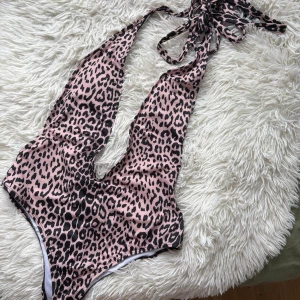 Leopardmönstrad rosa baddräkt med knyt - Snygg baddräkt i rosa med leopardmönster och djup urringning. Modellen har knyt i nacken för en justerbar passform och är tillverkad i ett stretchigt syntetmaterial. Perfekt för strandhäng eller poolparty.