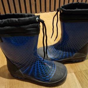 Gummistövlar med lätt fodring och snörning - Säljer ett par blå stövlar med svart prickigt mönster och svarta detaljer. Stövlarna har snörning upptill för att justera passformen och en robust sula som passar perfekt för regniga dagar.