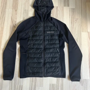 Svart pufferjacka Peak Performance S - Mörk blå cardigan från Peak Performance med huva och hel dragkedja. Jackan har quiltade partier fram, bak och på huvan, samt släta ärmar. Två dragkedjeförsedda fickor framtill. Perfekt för dig som gillar sportig och clean stil.