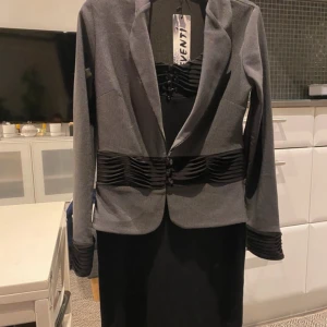 Grå och svart kavaj från Eventi - Snygg grå kavaj från Eventi med svarta veckade detaljer i midjan och vid ärmsluten. Kavajen har klassisk krage, knäppning framtill och en figurnära passform. Perfekt för dig som gillar stilrena och unika plagg med coola detaljer.