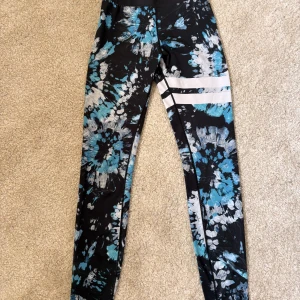 Stronger träningsbyxor - Snygga leggings med svart bas och blått samt vitt batikmönster. Två vita ränder på ena benet ger en sportig vibe. Perfekta för träning eller chill. Stretchigt material som sitter tajt och bekvämt.