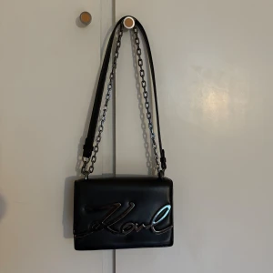 Svart axelväska från Karl Lagerfeld - Snygg svart axelväska från Karl Lagerfeld i slätt skinn med metallkedja och stor silverfärgad Karl-logo framtill. Väskan har en stilren, rektangulär form och coola detaljer som ger en modern vibe. Väskan har även defekter på loggan som finns på bild 3, så kom gärna med egna prisförslag oxå☺️🙌🏽