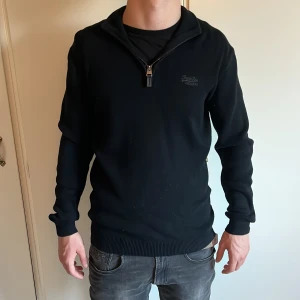 Svart half zip-tröja från Superdry - Svart långärmad tröja från Superdry med half zip-dragkedja vid halsen. Tröjan är i mjuk bomullsblandning med 5% kashmir för extra komfort. Ribbstickade muddar och diskret logga på bröstet. Perfekt för lager-på-lager-stil. Modellen är 178 cm lång och väger 64 kg. Det är bara att höra av sig vid frågor och funderingar 😀