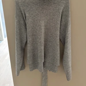 Grå stickad polotröja från Pieces - Mysig grå stickad polotröja från Pieces med långa ärmar och ribbade muddar. Tröjan har en hög polokrage och snygg knytning i ryggen som ger en unik touch (går att knyta runt midjan). Perfekt för kyliga dagar nu till hösten/vintern och enkel att styla med jeans eller kjol.