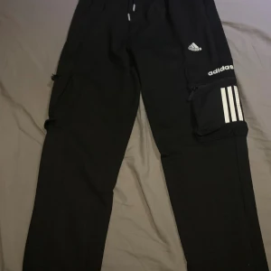 Svarta Adidas mjukisbyxor med dragkedja - Svarta mjukisbyxor från Adidas med vita loggor och klassiska tre ränder på benet. Byxorna har snörning i midjan och en praktisk ficka med dragkedja på sidan. Perfekta för chill eller träning, riktigt sköna och snygga.De finns adidas märke i både sidorna 