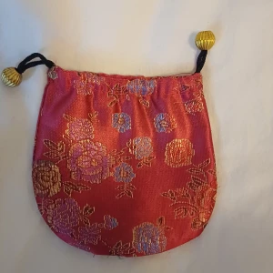 Röd blommig sidenpåse med snörning - Supersöt liten röd påse i siden med glansigt blommönster i guld och lila. Stängs med svart snörning och har dekorativa guldfärgade pärlor i ändarna. Perfekt för småprylar eller smycken.