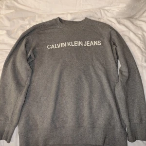 Grå Calvin Klein Jeans sweatshirt S - Säljer en stilren grå sweatshirt från Calvin Klein Jeans i storlek S. Tröjan har rund halsringning, långa ärmar och vit logotyp framtill. Perfekt basic med ribbade muddar och mjukt material som passar till allt.