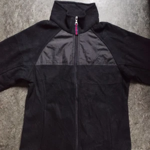 Tommy Hilfiger Fleecejacka - Svart fleecejacka från Tommy Hilfiger med dragkedja framtill, hög krage och snygga detaljer i nylon över bröstet. Jackan har snörning i nederkant och blåvitrandigt innerfoder i fickorna. Perfekt för kyliga dagar och enkel att matcha.