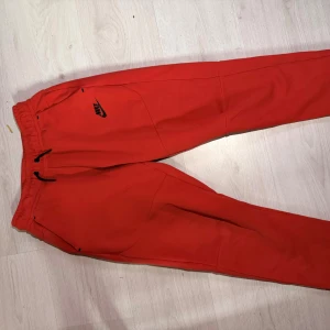 Röda mjukisbyxor från Nike - Riktigt snygga röda mjukisbyxor från Nike med svart logga på framsidan och svarta dragsnören i midjan. Byxorna har en dragkedjeficka bak och är tillverkade i mjukt bomullsmaterial. Perfekta för chill eller träning. Priset ej hugget