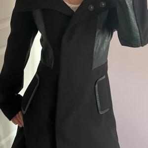 Svart kappa med skinnpaneler - Säljer en svart kappa med snygga skinnpaneler på axlar och fickor. Jackan har stora knappar framtill och en stilren, lite oversized passform. Perfekt för dig som gillar en edgy och modern look.