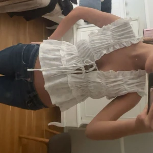 Vit blommig blus med volang från Gina Tricot - Supersöt vit blus från Gina Tricot med ljusblått blommönster, volangkant och knytning framtill. Modellen är ärmlös och har en croppad passform med romantiska detaljer. Perfekt för en fräsch och somrig look!