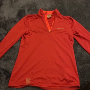 Röd långärmad träningströja Craft - Sportig röd långärmad träningströja från Craft i funktionsmaterial. Tröjan har halv dragkedja framtill, hög krage och diskret logga på bröstet. Perfekt för löpning eller gym och har en snygg, enkel design med orange detaljer vid dragkedjan.