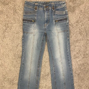 Blå jeans med dragkedjedetaljer - Snygga ljusblå jeans med raka/bootcut ben och coola dragkedjor framtill. Flera fickor både fram och bak med knappdetaljer. Jeansen har en klassisk passform och markerade sömmar som ger en schysst look.  Midja rakt över: 35cm Lår: 22cm Innerben:73cm