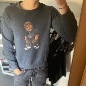 Mörkgrå sweatshirt med nalletryck - Säljer en mörkgrå sweatshirt med ett coolt tryck av en streetwear-nalle på bröstet. Tröjan har rund hals och långa ärmar, perfekt för dig som gillar avslappnad stil med en twist. Passar till jeans eller joggers för en chill look.