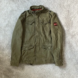 Superdry Fieldjacka militärgrön - Hej! Militärgrön fieldjacka från märket Superdry säljes! Jackan är i ett gott skick utan defekter. Jackan har 4st praktiska fickor, snygga detaljer och en brittisk patch i läder på armen. Perfekt för kyligare dagar samt håller dig unik och stilren. Jackan är storlek XS och som referens så är jag 180cm och väger ca 67kg, rekommenderar att vara mindre än det. Skriv vid minsta intresse 😊mvh DC🔹