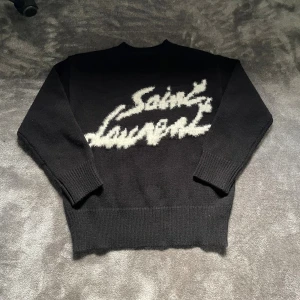 Svart Saint Laurent stickad tröja - Svart stickad tröja från Saint Laurent med vit, fluffig logga framtill. Tröjan har rund hals, långa ärmar och breda ribbade muddar vid ärmslut och nederkant. Perfekt för dig som vill ha en statement piece med lyxig känsla.
