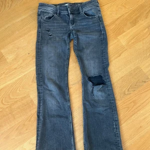 Grå slitna jeans med hål - Säljer ett par grå jeans med coola slitningar och hål på låret och vid fickan. Bootcut och låg midja. Jeansen har en snygg tvättad look och är perfekta för en avslappnad streetstil.