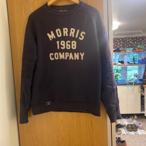 Mörkblå stickad tröja Morris Gents - Snygg mörkblå stickad tröja från Morris Gents med broderad text 'Morris 1968 Company' framtill. Tröjan har ribbade muddar vid ärmslut och nederkant samt klassisk rund hals. Perfekt för dig som gillar stilren och sportig look.