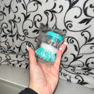 Diskborste tvåldispenser  - Bilderna visar en handhållen diskborste med inbyggd tvåldispenser, ny i förpackning. Borsten har turkosa borst och ett transparent handtag med vit och turkos topp. Perfekt för effektiv rengöring av disk och köksredskap. Inga synliga defekter eller slitage.