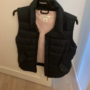 Svart dunväst från H&M L.O.G.G. - Svart dunväst från H&M L.O.G.G. med quiltad design och hög krage. Västen har dragkedja framtill och två fickor med lock och knappdetalj. Perfekt att slänga över en hoodie för en chill look.