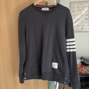 Mörkgrå sweatshirt från Thom Browne - Säljer en mörkgrå sweatshirt från Thom Browne med klassiska vita ränder på ena ärmen och märkets etikett framtill. Tröjan har rund halsringning och ribbade muddar. Perfekt för en stilren och sportig look.