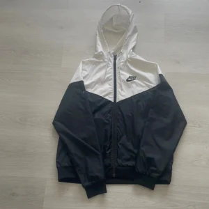 Nike svartvit vindjacka med huva L - Nike vindjacka i svart och vitt med huva och hel dragkedja framtill. Jackan har klassisk sportig look, elastiska muddar och meshfoder. Snygg tvåfärgad design med Nike-logga på bröstet och detaljerad dragkedja.