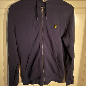 Mörkblå hoodie från Lyle & Scott - Mörkblå hoodie från Lyle & Scott med dragkedja framtill och klassisk huva. Tröjan har två fickor och den ikoniska gula loggan broderad på bröstet. Perfekt för chill dagar eller när du vill ha en enkel men snygg look.
