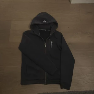 Svart hoodie från Sail Racing - Svart hoodie från Sail Racing med dragkedja framtill och broderad logga på bröstet. Tröjan har huva med tryckt detalj och långa ärmar. Perfekt för dig som gillar stilren och sportig look.
