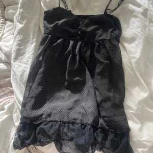 Svart polkadot kortklänning med volang - Svart kortklänning/top med tunna axelband, små vita prickar och dekorativa knappar framtill. Kjolen har ett lager av transparent volang längst ner som ger en extra touch. Perfekt för dig som gillar en söt och lekfull stil. ”Vintage h&m”🥰🥰